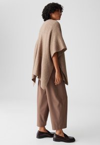 Beige gebreide poncho met een losse silhoutte, V-vormige zoom en geribbelde textuur, gecombineerd met bruine wijde broek en donkere schoenen.