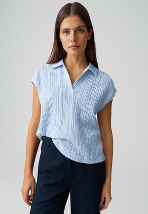 Young woman wearing a light blue textured short-sleeve top with collar and black high-waisted pants, standing against a plain background.