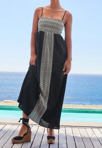 Femme portant une robe maxi noire avec un motif géométrique blanc, se tenant sur une terrasse en bois près de la piscine et de l'océan, chaussée de sandales compensées.