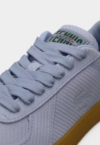 Baskets en velours côtelé bleu clair avec surface texturée, lacets assortis, étiquette logo verte et semelle en gomme. Présente un détail de logo subtilement embossé.