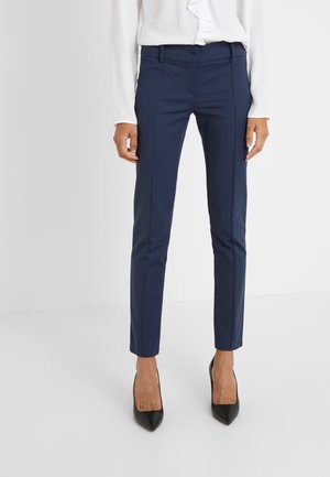Femme portant un pantalon slim bleu marine et des escarpins noirs à bout pointu, assortis à un chemisier blanc à volants, debout sur un fond uni.