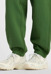 Grüne Jogginghosen mit elastischen Bündchen, kombiniert mit beigen Mesh-Sneakern mit weißen Schnürsenkeln und dezenten Akzenten. Weiche, legere Stoffstruktur.