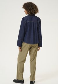 Giacca di jeans in blu scuro con maniche lunghe, presenta dettagli di cucitura. Abbinata a pantaloni beige chiaro e scarpe nere. Vista posteriore mostrata.