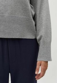 Maglione grigio a maglia con polsini a costine, abbinato a pantaloni blu scuro. La vista ravvicinata mette in evidenza la morbida texture e i colori a contrasto.