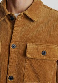 Cămașă din corduroy maro cu nasturi, prezentând o textură ribbed, nasturi negri și buzunare pe piept cu clapete. Vedere de aproape a gulerului și a materialului.