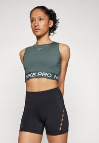Nike Pro športni modrček v temno zeleni barvi z belim logotipom, skupaj z visokim črnim šortsom z stranskimi izrezi. Gladek material, prilegajoča se zasnova.
