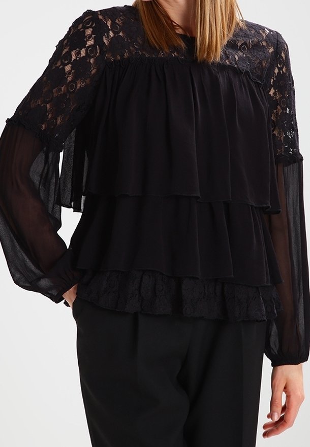Blouse noire à volants avec des accents en dentelle sur les manches et le décolleté. Tissu transparent avec des couches froncées et une coupe décontractée.