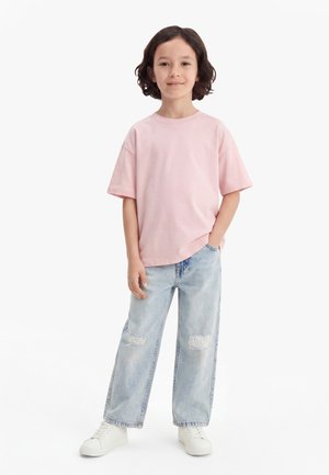 Dziecko o ramionowych kręconych włosach, ubrane w jasnoróżowy, oversize'owy T-shirt, szerokie jasnoniebieskie jeansy z łatkami w kształcie misiów oraz białe trampki.