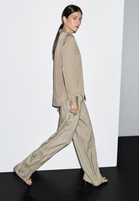 Beige blouse met lange mouwen en wijde, geruite broek. Soepele stof met een ontspannen pasvorm. Model loopt tegen een minimale achtergrond.