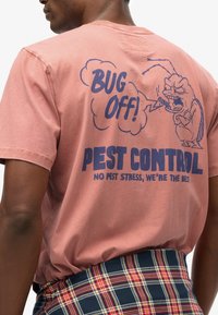 Camiseta de algodón rosa con un bicho de dibujos animados, texto "¡VETE, BICHO!" y "CONTROL DE PLAGAS" en azul. La camiseta tiene un corte holgado y un diseño de cuello redondo.