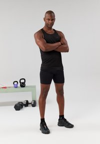 Camisola de desporto preta e calções usados por um homem em pé, com sapatilhas pretas. Kettlebells e halteres sobre uma mesa de vidro próxima.
