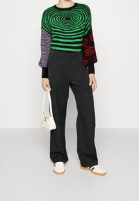 Pull multicolore avec motif en spirale verte, manches violettes et noires; pantalon noir taille haute; sac blanc et baskets avec rayures noires.