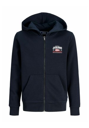Garçon blond debout, portant un sweat à capuche noir zippé, un t-shirt blanc, un jean bleu ample et des baskets blanches, sur un fond clair uni.