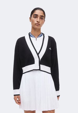 Femme aux cheveux tirés porte un cardigan court noir avec bordure blanche sur une robe plissée blanche avec col rayé bleu sur fond clair.