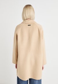 Abrigo de lana beige con un corte relajado, mangas raglán y un cuello redondo. La parte posterior presenta una pequeña etiqueta de marca negra. Textura suave.