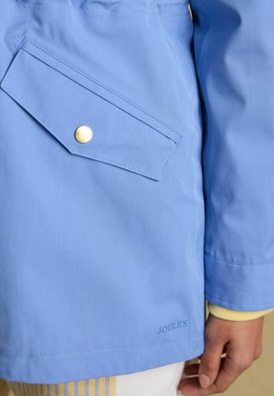 Svetlomodrý rukáv bundy a vrecko so zlatým patentom, čiastočný pohľad na ruku a žltý pruhovaný odev pod ním.