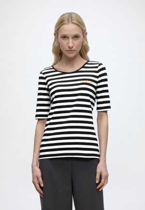 Vrouw met lang blond haar draagt een zwart-wit horizontaal gestreept shirt met korte mouwen en donkere broek, staand tegen een effen achtergrond.