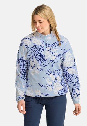 Frau mit blondem Zopf, die eine hellblaue gemusterte Fleecejacke mit Viertelreißverschluss und dunkelblaue Hose trägt, schaut nach unten und zur Seite.