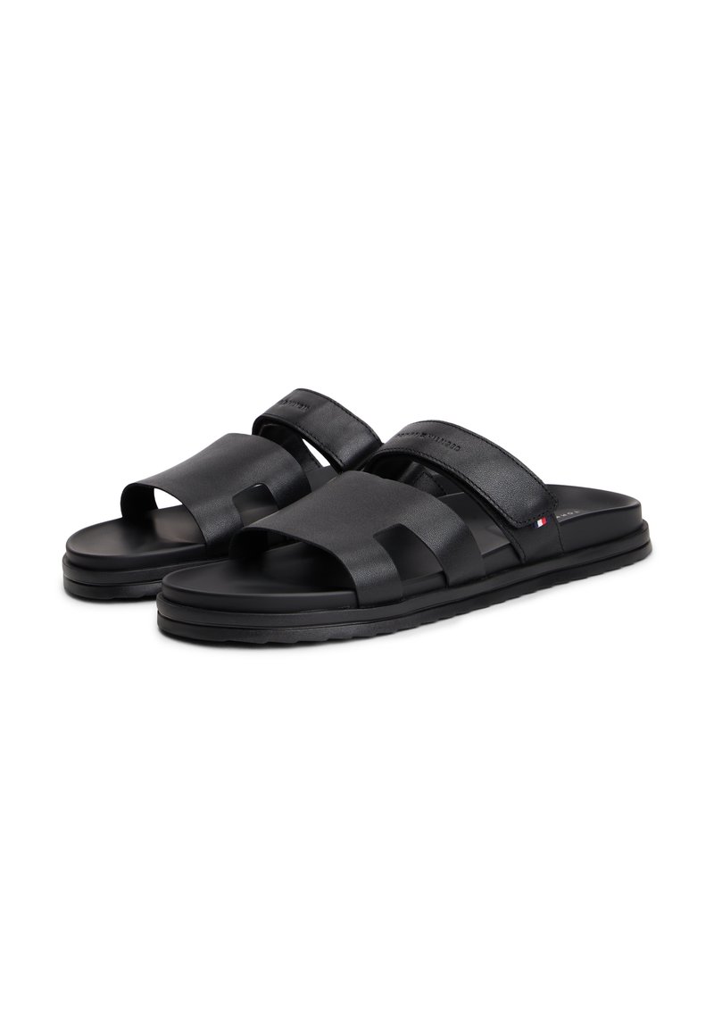 Sandalias negras tipo slide con dos correas gruesas, textura suave y suela acolchada. Presentan bordes curvos y una marca minimalista en rojo, blanco y azul.