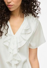 White blouse featuring ruffled front detail, short sleeves, and a V-neckline made of lightweight, flowing fabric.