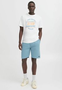 Tricou alb cu text grafic din denim, asociat cu pantaloni scurți bleu deschis și teniși albi. Dressul are un design casual și o croială lejeră.