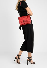 Punainen käärmekuvioinen käsilaukku, jossa on läppä ja pyöreät metalliosat, yhdistettynä mustaan haalariin ja mustiin niitattuihin korkokenkiin.