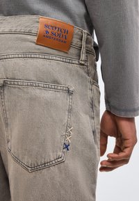 Ανοιχτό γκρι τζιν με ραφές στην πίσω τσέπη και καφέ δερμάτινο λογότυπο με την επιγραφή "Scotch & Soda Amsterdam", φορεμένα από άτομο με γκρι πουκάμισο.