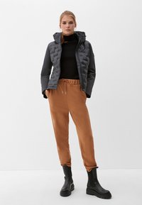 Grau gepuffte Jacke mit gestepptem Muster, schwarzer Rollkragenpullover und braune Jogginghose mit elastischem Bund, kombiniert mit schwarzen Stiefeletten.