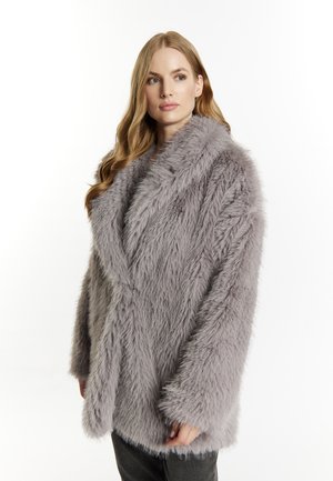 Femme aux longs cheveux blonds portant un manteau oversize en fausse fourrure gris clair sur fond blanc.