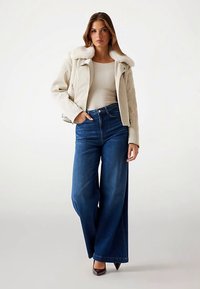 Beige jakke foret med kunstpels med lynlås foran, cremefarvet top og vidbenede mørkeblå jeans. Brune spidse hæle fuldender outfittet.