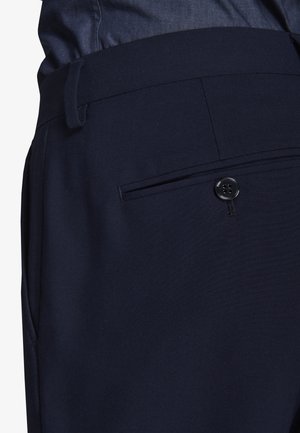 Nærbillede af marineblå bukser, der viser taljebånd med bæltestropper og baglomme sikret med en sort knap.