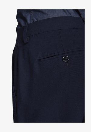 Nærbillede af marineblå bukser, der viser taljebånd med bæltestropper og baglomme sikret med en sort knap.