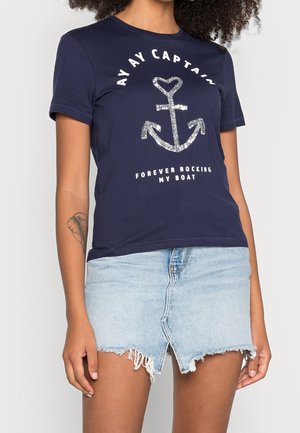 Kobieta ubrana w granatowy T-shirt z błyszczącym kotwicą i napisem "AY AY CAPTAIN FOREVER ROCKING MY BOAT" w połączeniu z jasną, postrzępioną dżinsową spódnicą.