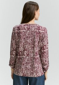 T-shirt à manches longues en bordeaux avec un motif abstrait texturé lumineux. Doté d'un col rond et d'une coupe légèrement ajustée. Le tissu semble doux.