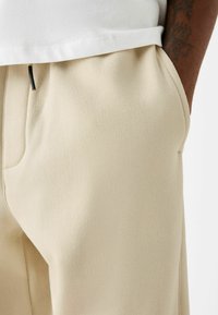 Närbild av en person som bär beiga mjukisbyxor med en sidficka, en vit skjorta och en synlig tatuering på armen.