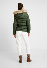 Zielona kurtka puchowa z kapturem z sztucznego futra, pikowana tekstura i dopasowany krój, w połączeniu z białymi spodniami i brązowymi botkami na obcasie w wężowy wzór.