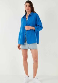 Blå skjorte med knaplukning i struktureret stof, to forlommer og en afslappet pasform, parret med en denimnederdel og hvide sneakers.