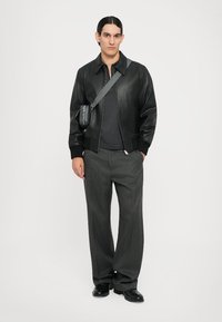 Veste aviateur en cuir noir avec des poignets côtelés, t-shirt gris en dessous, pantalon large gris et sac bandoulière. Design minimaliste.