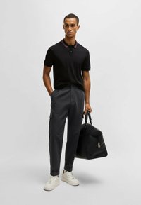 Svart pikétröja med burgundikrage, i kombination med grå randiga byxor. Modellen håller en svart duffelväska och bär vita sneakers.