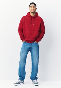 Rode hoodie met trekkoorden en voorzak, gecombineerd met lichtblauwe spijkerbroek en grijze sneakers. Effen achtergrond, model staat ontspannen.