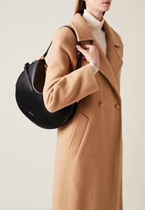 Borsa a tracolla in pelle nera con forma arrotondata, dettagli in metallo dorato e superficie liscia, abbinata a un cappotto color cammello.