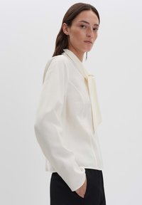 Blusa bianca con colletto a fiocco, maniche lunghe, texture liscia, vestibilità sartoriale e dettagli delle cuciture sottili. Abbinata a pantaloni scuri.