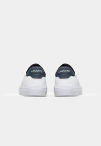 Hvite sneakers med en teksturert overdel, marineblå hælakkenter og kontrasterende hvite snørebånd. Har "LACOSTE"-logoen på hælen.