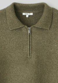 Pull-over à col zippé de couleur vert olive, fabriqué en tissu tricoté, avec une fermeture éclair argentée et une étiquette portant le nom de la marque et la taille. La texture est douce.
