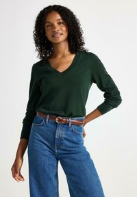Grön v-neck tröja i mjukt material, kombinerad med högmidjade blå jeans och ett brunt bälte med en silverbuckle.