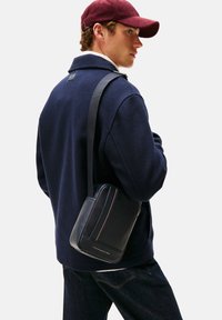 Musta nahkainen crossbody-laukku, jossa on teksturoitu pinta, kontrastitikkaukset ja merkitty edusta. Käytetty olkapäällä tummansinisen takin kanssa.