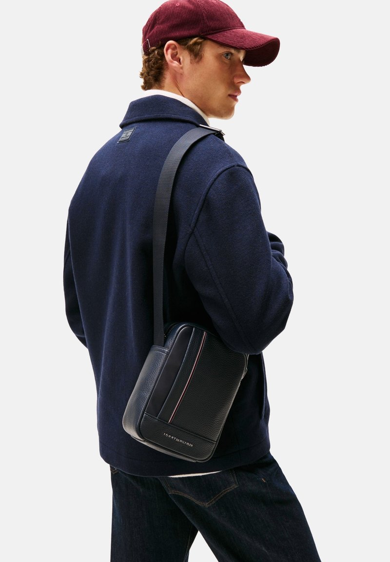 Musta nahkainen crossbody-laukku, jossa on teksturoitu pinta, kontrastitikkaukset ja merkitty edusta. Käytetty olkapäällä tummansinisen takin kanssa.