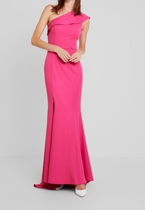 Femme en robe longue rose vif asymétrique avec une fente latérale, portant des escarpins blancs pointus, debout devant un fond uni.