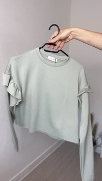Felpa cropped verde chiaro con maniche lunghe e dettagli arricciati sulle spalle, realizzata in tessuto morbido e liscio. Presenta un collo rotondo.