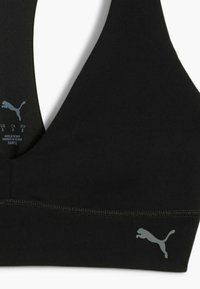 Crna sportska grudnjak s niskim izrezom i sivim Puma logotipom s lijeve strane. Oznaka za njegu vidljiva unutar odjeće.